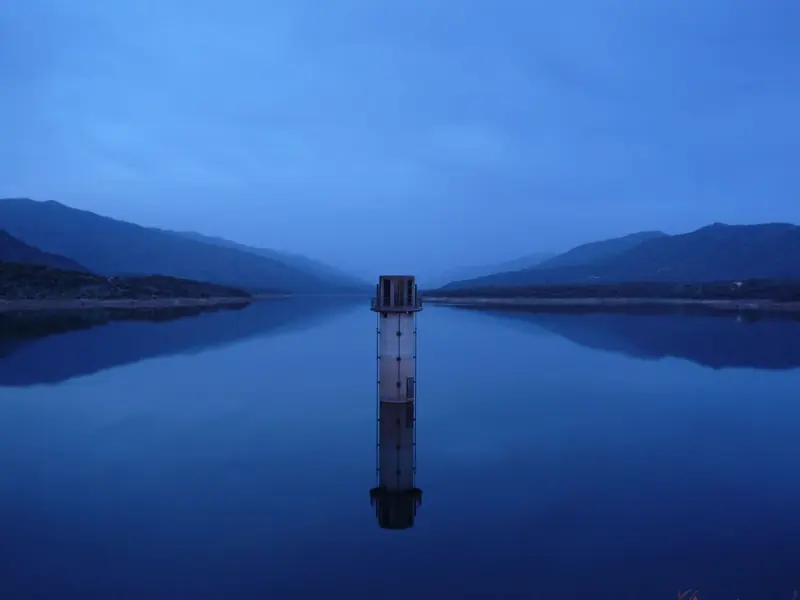 Torre de toma de un embalse reflejada en el agua al anochecer, entre montañas azules