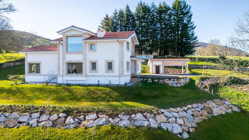 Casa blanca sobre un prado con muro de piedra y pinos al fondo, en el valle de Mazcuerras